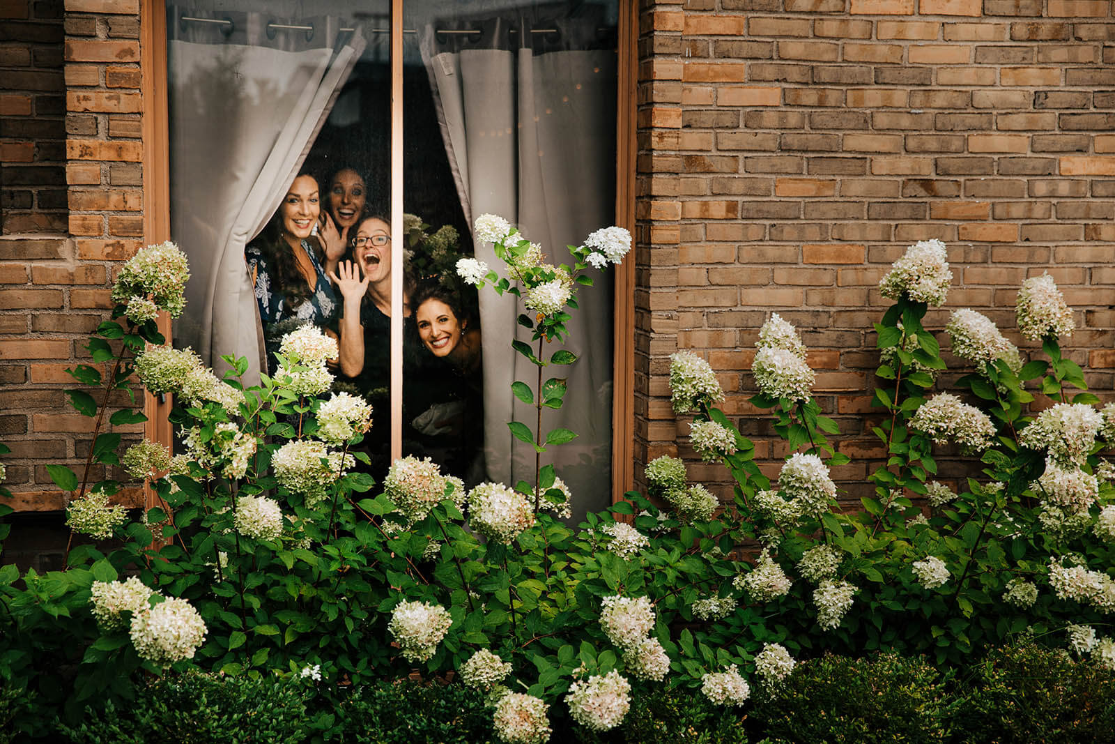 Kensington Hotel wedding photographer Ann Arbor Michigan candid bridesmaids peeking and waving through window surrounded by hydrangeas getting ready, top Michigan luxury wedding photography