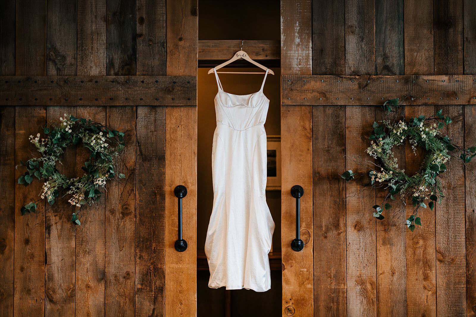 Wedding dress hanging on rustic barn doors with greenery wreaths at Westers Family Winery in Junction Michigan