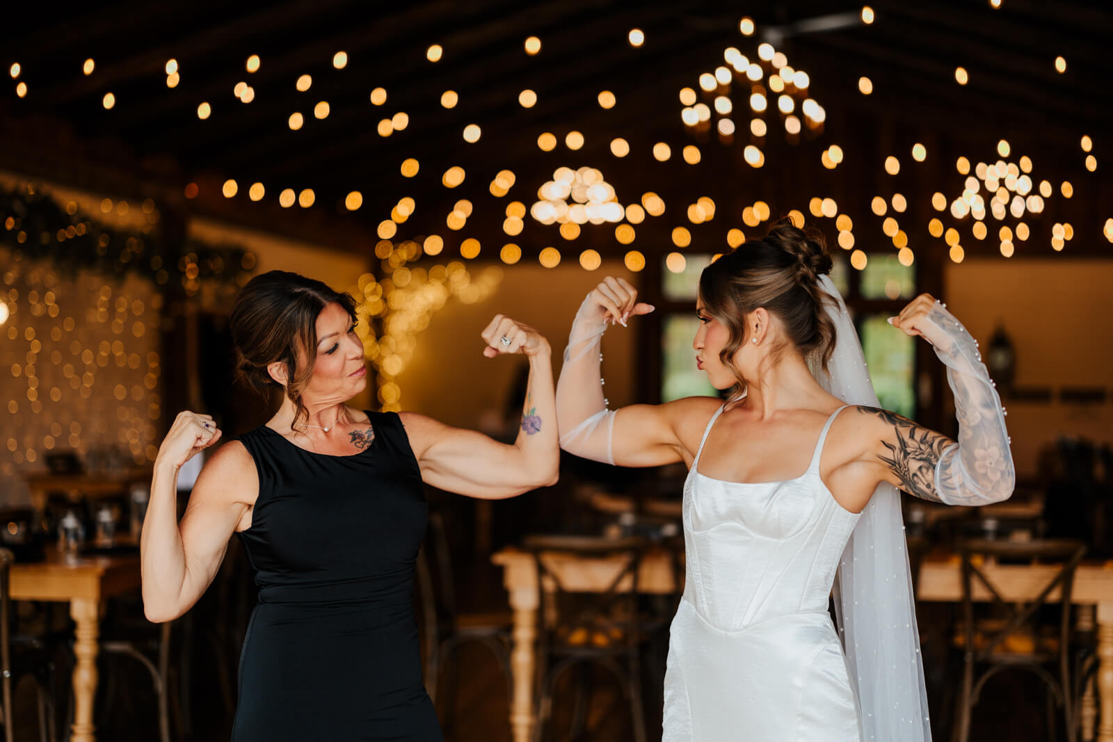 Bride and mother flexing muscles together and laughing under string lights