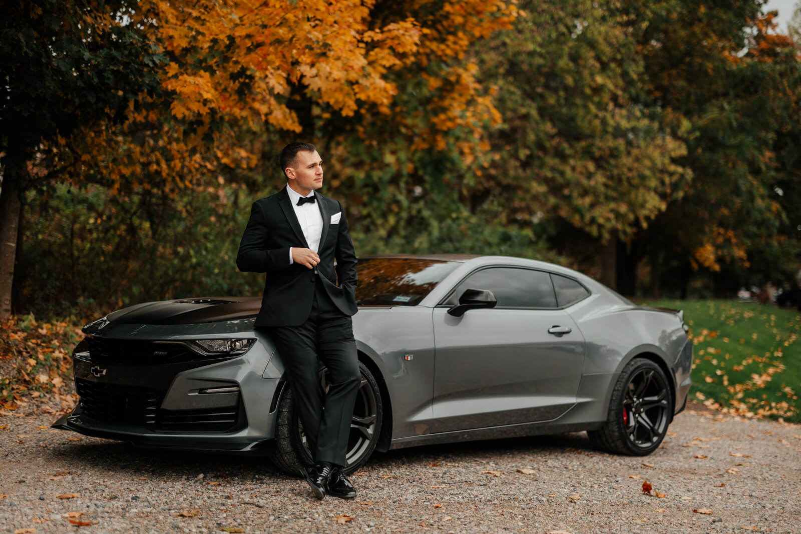 Groom in black tuxedo leaning against gray Camaro at fall winery wedding