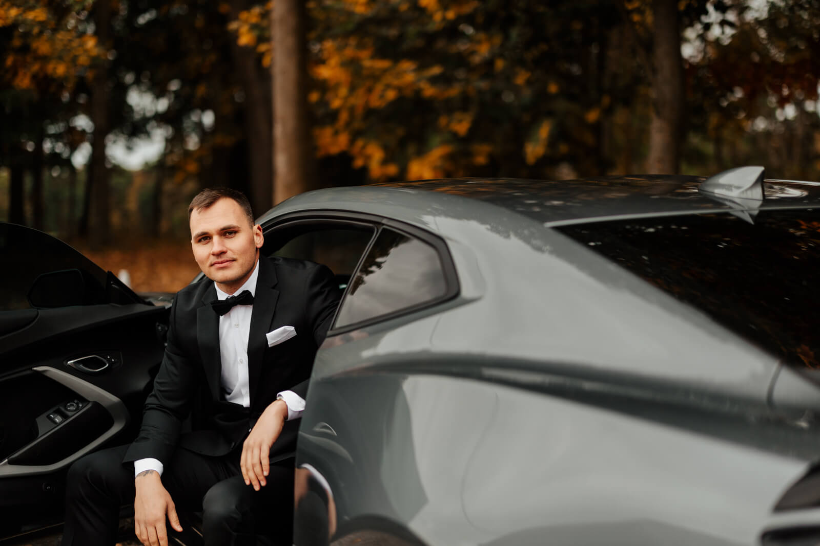 Groom sitting in open door of gray Camaro in tuxedo at October wedding