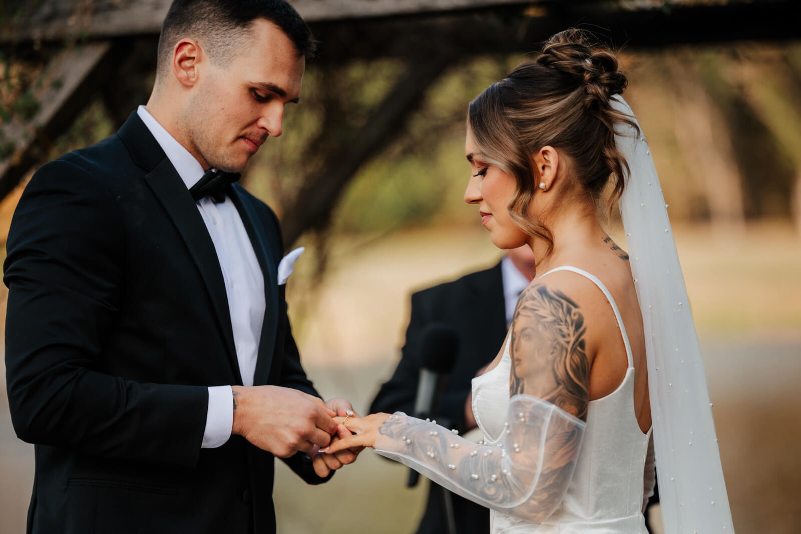 Groom placing wedding ring on bride's finger during outdoor ceremony