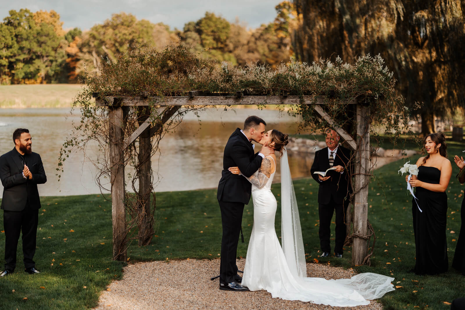 Bride and groom first kiss under wooden pergola arch with pond behind at Westers Family Winery