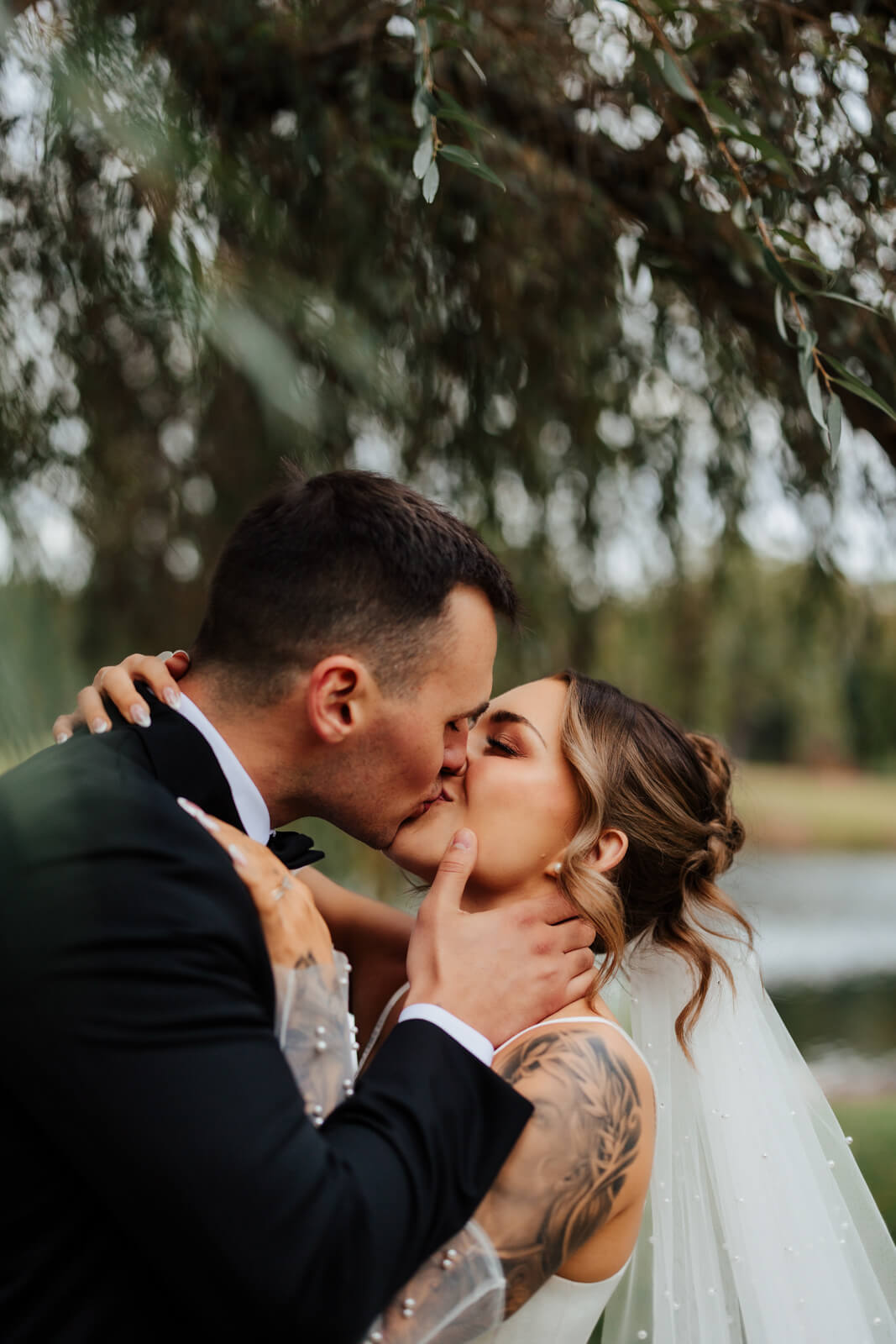 Groom kissing bride's cheek under weeping willow tree at Westers Family Winery