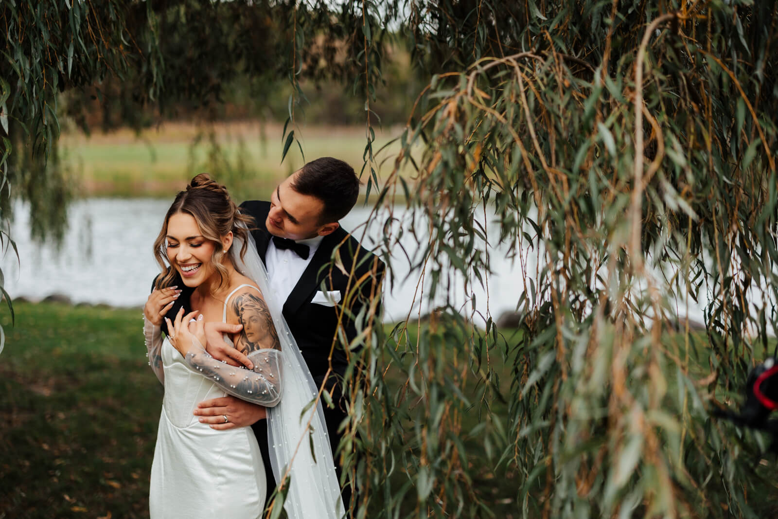 Bride and groom laughing together under weeping willow by pond at Westers Family Winery