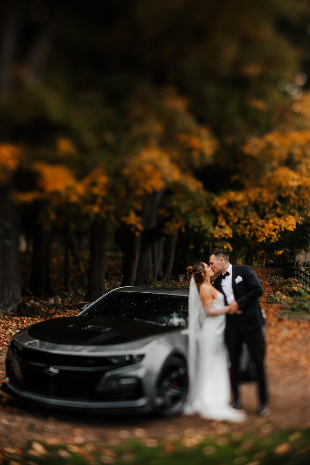 Bride and groom kissing in front of Camaro with fall trees at Westers Family Winery