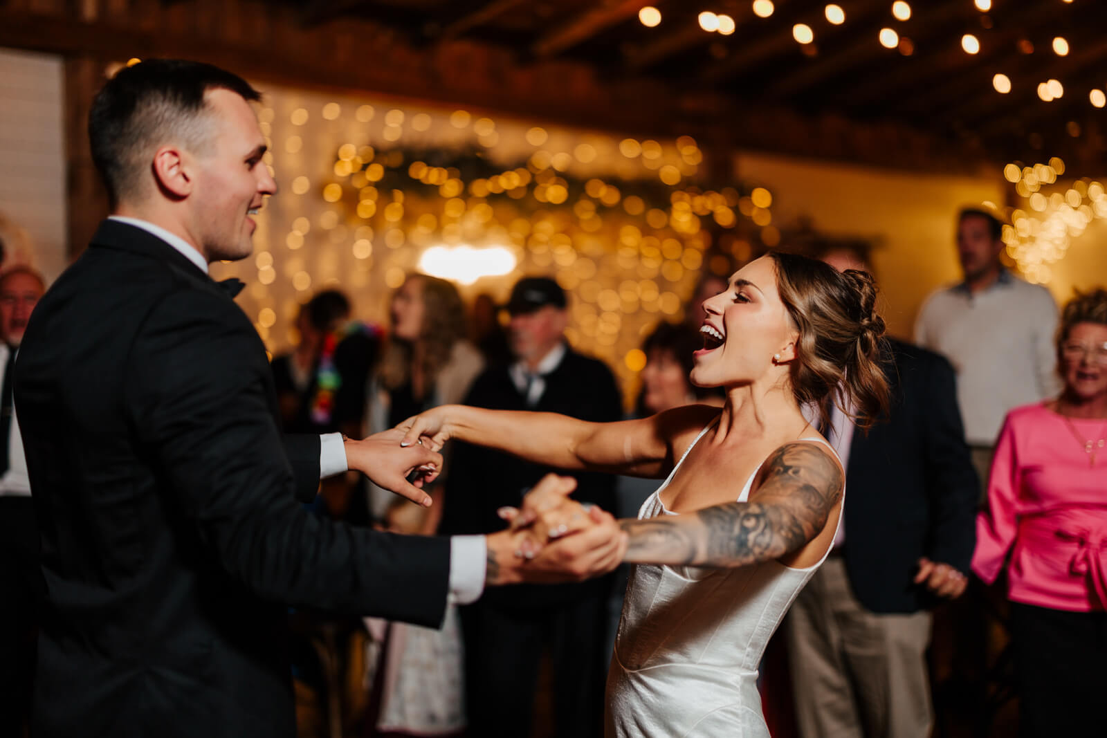 Bride laughing and dancing with groom on packed dance floor under string lights at winery reception