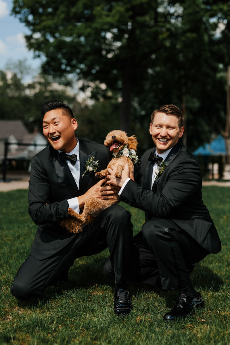 Same-sex couple in tuxedos laughing with their doodle dog wearing floral collar at Grand Rapids wedding
