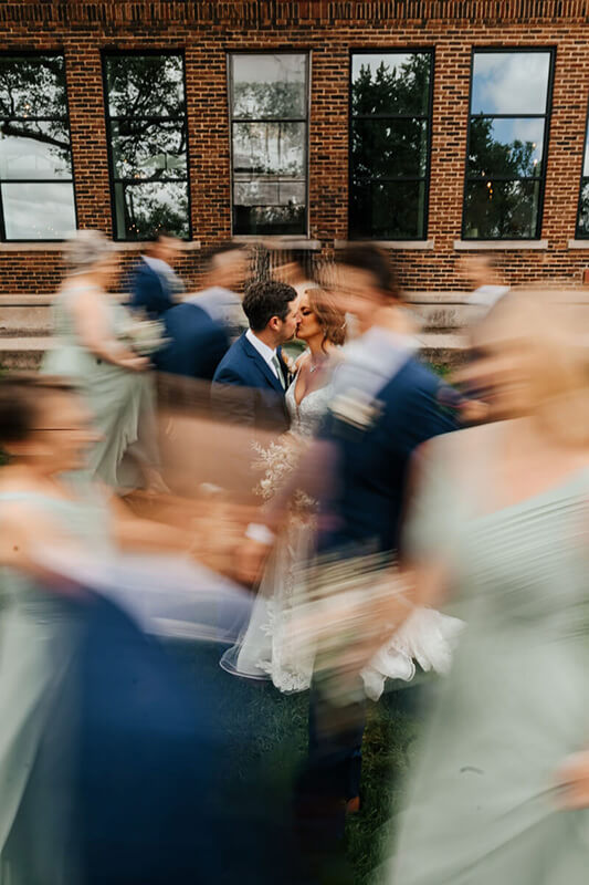Motion blur bridal party surrounds kissing bride and groom outside brick building at Grand Rapids wedding