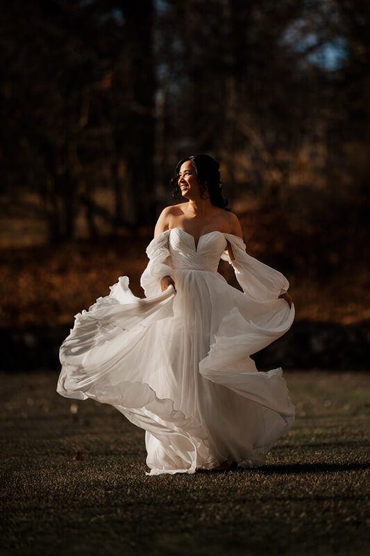 Bride spinning in flowing off-shoulder gown at golden hour with dark wooded background