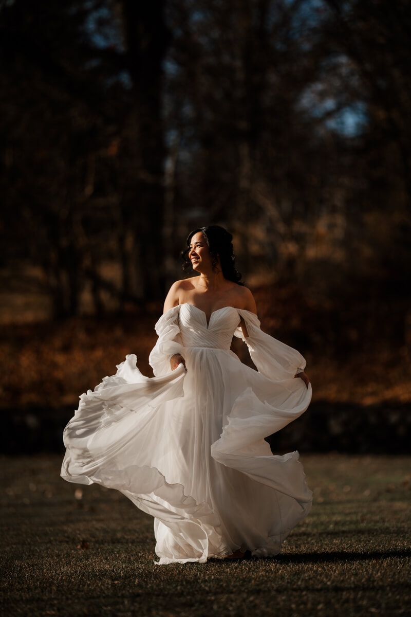 Bride spinning in flowing off-shoulder gown at golden hour with dark wooded background