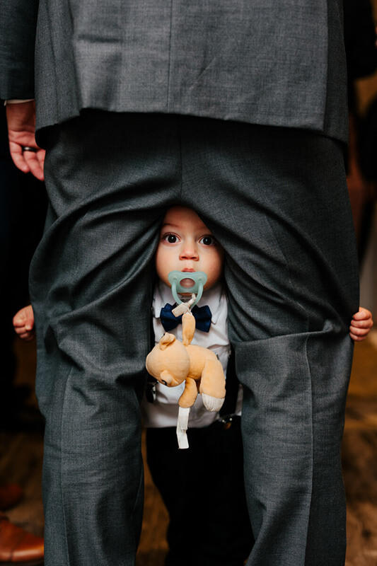 Adorable baby in bow tie peeking through dad's suit jacket holding stuffed animal with pacifier