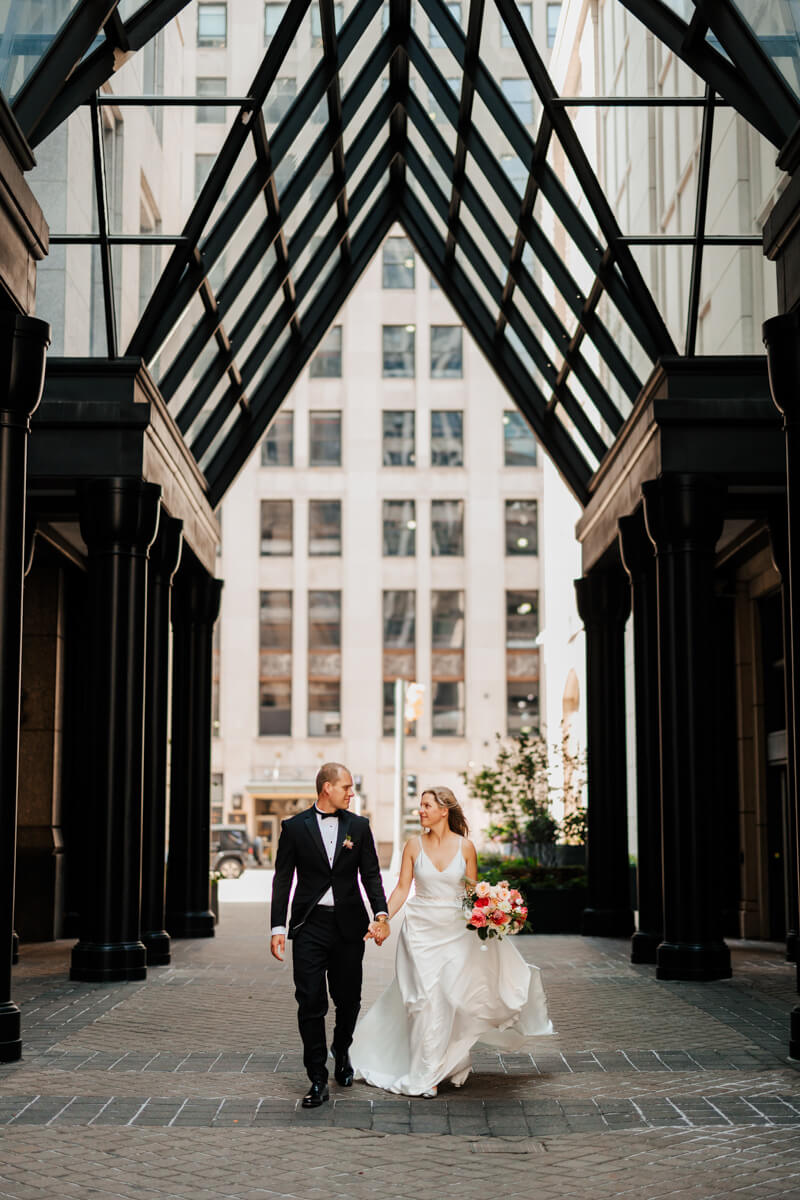Bride and groom walking hand in hand under glass canopy archway in downtown Grand Rapids